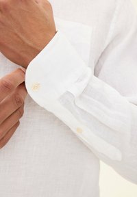 Camicia bianca di lino con polsini abbottonati. Il tessuto appare testurizzato e leggero. Il polsino ha due bottoni ed è leggermente arrotolato.