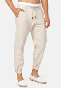 Man in beige casual pants met een touwtje in de taille en opgerolde zoom, gecombineerd met witte instap schoenen op een witte achtergrond.