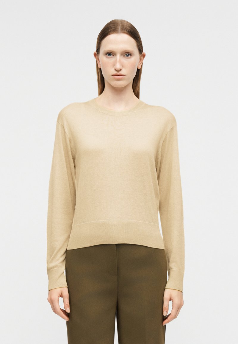 Femme aux cheveux bruns et raides porte un pull à manches longues beige et un pantalon vert olive, debout devant un fond clair uni.