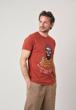 Homme avec barbe portant un t-shirt couleur rouille avec un graphique d'homme barbu en lunettes de soleil et un oiseau, debout les mains dans les poches, regardant vers la droite.