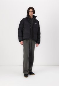 Svart boblejakke med glidelås foran, elastiske mansjetter og sidelommer. Båret over en mørk skjorte og kombinert med grå bukser og svarte joggesko.