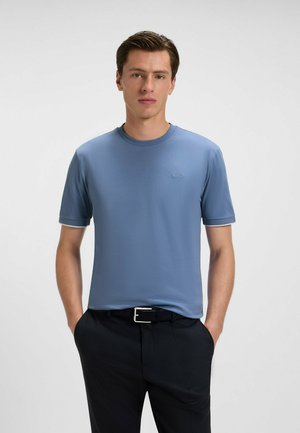 T-shirt azzurro chiaro a maniche corte con collo rotondo e un lieve dettaglio del logo. Texture liscia, abbinato a pantaloni neri e una cintura.
