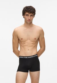 Czarne bawełniane bokserki o jednolitym wzornictwie, z elastycznym paskiem w logo i białym napisem. Noszone przez modela w neutralnej pozie.