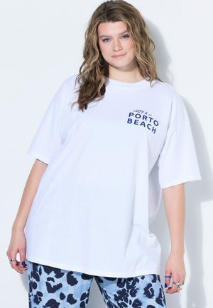 Ung kvinde med bølget hår iført en hvid oversized T-shirt med teksten "PORTO BEACH" og blå mønstrede bukser, poserer foran en hvid baggrund.
