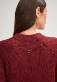 Bordeaux gebreide trui met een gestructureerd oppervlak, met een ronde halslijn en een klein metalen logo op de achterkant.