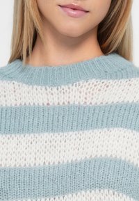 Nærbilde av en persons nedre ansikt og hals som har på seg en lukseriøs, blå og hvit stripete strikkegenser.