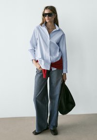 Blau-weiß gestreiftes Hemd mit Knöpfen, roter Pullover in der Taille gebunden, weit geschnittene blaue Jeans, schwarze Handtasche und große schwarze Sonnenbrille.