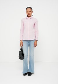 Camisa de botão rosa e branca com riscas, combinada com jeans claros de boca de sino. Uma mala preta e sapatos completam o visual.