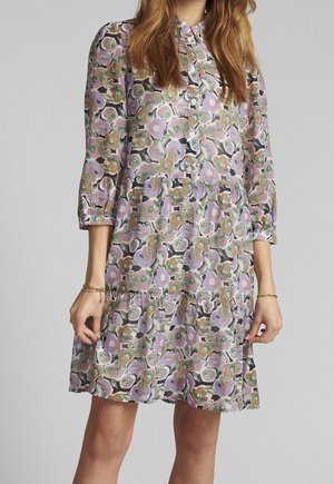 Woman wearing a buttoned floral dress with purple, green, and brown patterns, 3/4 sleeves, and ruffled tiers above the knee.