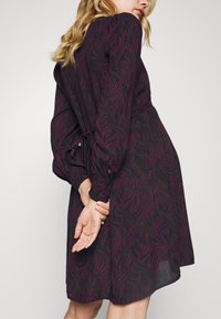 Femme portant une robe noire à manches longues avec des motifs abstraits de feuilles violettes, debout les mains jointes dans le dos.