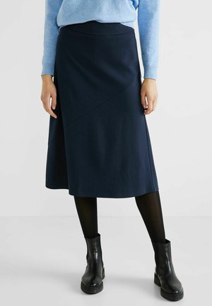 Woman wearing a navy midi skirt, black tights, black ankle boots, and a light blue sweater, standing against a plain background.