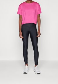Person som bär en rosa kortärmad t-shirt, högmidjade svarta leggings och vita sportskor, stående mot en enfärgad bakgrund.