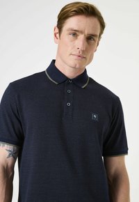 Polo shirt bleu marine avec un motif texturé subtil, un col contrastant et une patte de boutonnage à trois boutons. Présente un petit logo sur la poitrine.