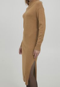 Vestido de punto en color camel, con cuello alto, mangas largas y aberturas laterales. Textura suave y silueta ajustada.