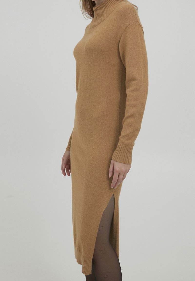 Vestido de punto en color camel, con cuello alto, mangas largas y aberturas laterales. Textura suave y silueta ajustada.