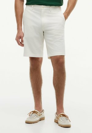 Mann trägt weiße maßgeschneiderte Shorts, weiße geflochtene Lederschuhe mit braunen Schnürsenkeln und ein grünes Hemd, das in die Hose gesteckt ist, steht mit einer Hand in der Hosentasche.