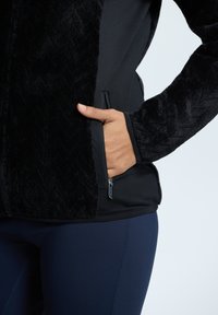 Giacca nera con zip, con pelliccia sintetica testurizzata sul davanti e tessuto liscio sui lati. Presenta tasche laterali e un design aderente.