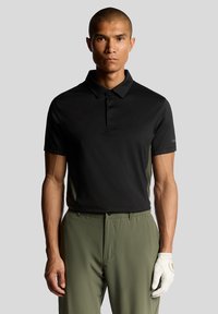 Sort polo-shirt med krave, knapper og kortærmede; parret med lys grønne bukser. En golfhandske er på venstre hånd.