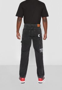 Man i svarta byxor med vitt logotyp och text på bakfickorna och röda, vita och svarta sneakers, stående mot en enkel ljus bakgrund.