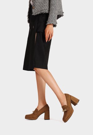 Mocassini a tacco a blocco in suede marrone con punta quadrata e dettagli in metallo dorato. Le scarpe hanno una texture liscia e una silhouette moderna.
