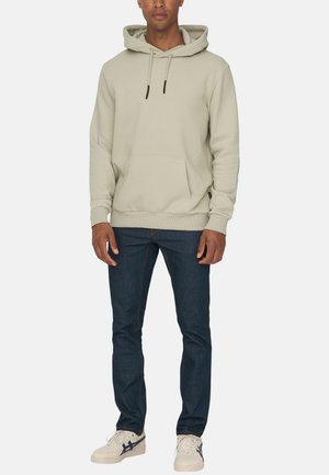 Beige hoodie met trekkoorden en een voorzak. Gepaard met donkerblauwe jeans en witte sneakers. Gladde stofstructuur.