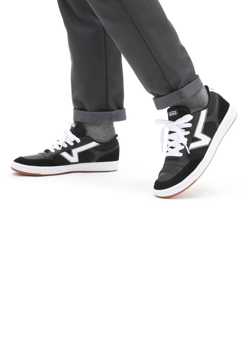 Svart og grå sneakers med hvite detaljer, med lavt design, snøring og en teksturert overflate, kombinert med grå bukser med oppbrett.