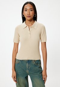 Beige geribde polo met een omgeslagen kraag, korte mouwen en vijf knopen. Draagt het met vervaagde blauwe wijde pijpen jeans.