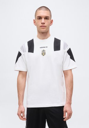 Man with short hair wearing white Adidas T-shirt with black stripes and Juventus football club logo, standing against plain background.