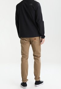 Schwarze Jacke mit Logo auf dem Rücken, khakifarbene Hose mit einem Patch und schwarze Turnschuhe. Das Outfit hat eine lässige Passform und besteht aus robusten Materialien.