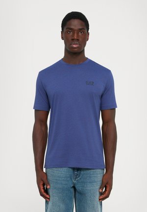 En svart mann som står med ansiktet fremover, iført en enkel blå EA7 T-skjorte og lyseblå jeans mot en hvit bakgrunn.