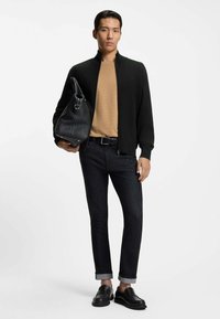 Mężczyzna ubrany w czarną kurtkę na zamek, beżową koszulę, ciemne podwinięte dżinsy, czarne mokasyny, trzymający dużą czarną skórzaną torbę na białym tle.