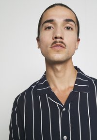 Marineblau gestreiftes Kurzarmhemd mit weißen vertikalen Streifen, leichtem Stoff und einem gekerbten Kragen. Verfügt über vordere Knöpfe und eine lockere Passform.