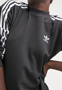 Svart sweatshirt med vit tre-randsdetaljering på ärmarna och en liten vit Adidas-logotyp på bröstet. Mjuk tygtextur.