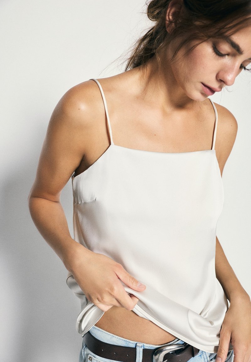 Femme en caraco blanc en satin, le levant légèrement pour révéler un jean bleu clair et une ceinture noire, regardant vers le bas, les cheveux attachés.