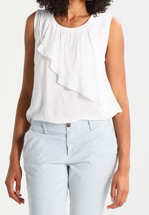 Ärmellose weiße Bluse mit Rüschen vorne, kombiniert mit hellblauen und weißen vertikal gestreiften Hosen, getragen von einer Person mit lockigem Haar.