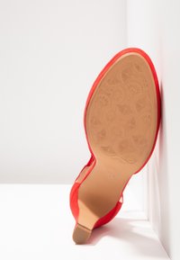 Chaussure à talon haut en daim rouge, positionnée avec la semelle vers le haut, montrant une semelle beige avec un motif floral embossé et un talon carré bas sur fond blanc.