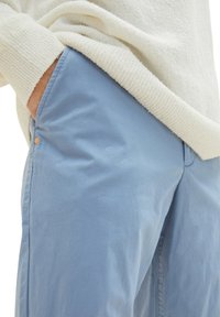 Jasnoniebieskie bawełniane spodnie o gładkiej teksturze i subtelnym blasku. Posiadają kieszeń boczną i mały pomarańczowy akcent w pobliżu pasa.