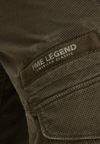 Tamnozeleni tkanina s teksturiranim uzorkom. Sadrži oznaku sa zakrpanom s natpisima "PME Legend" i "American Classic" u srebrnom slovu.