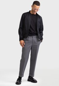 Blouson noir, t-shirt noir ajusté, pantalons gris à motifs et mocassins noirs. Le blouson présente un col et des poignets côtelés.