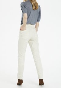 Pantalones ajustados beige con cintura alta, dos bolsillos traseros y detalles de costura sutiles. Combinados con una blusa azul de manga corta y tacones marrones.