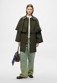 Casaco impermeável verde-oliva com design de capa em camadas, ferragens douradas e fecho de correr. Combinado com jeans de perna larga verde-claro.