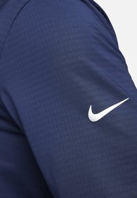 Tengeri kék atlétikai ing anyaga finom textúrával, bal ujján jól látható fehér Nike logóval.