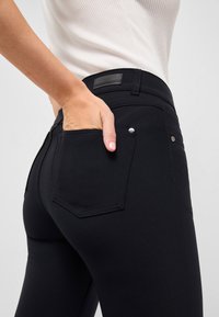 Schwarze Stretchjeans mit glatter Textur, ausgestattet mit Gesäßtaschen, sichtbaren Nähten und einem kleinen silbernen Knopfdetail in der Nähe der Tasche.