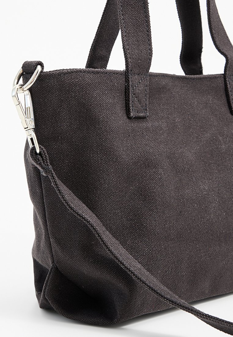 Bolso tote de tela negra texturizada con dos asas cortas y una correa desmontable sujeta por un clasp metálico plateado sobre un fondo blanco.