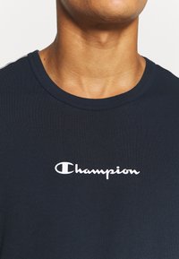 Námořnická modrá tričko s krátkým rukávem vyrobené z bavlny, s kulatým výstřihem, na hrudi s bílým logem "Champion". Hladká textura.