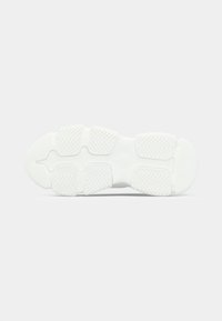 Hvit sneakersåle med segmenterte gummigrepputer som har fiskebensmønster, vist fra bunnsynsvinkel mot en ensfarget bakgrunn.