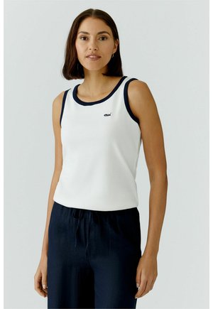 Femme aux cheveux bruns raides portant un haut blanc sans manches avec des bordures noires et un pantalon bleu marine, sur un fond clair uni.