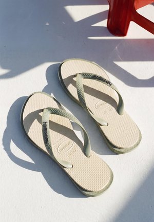 Paar beige Flip-Flops mit strukturierten Riemen und Sohlen, auf einer weißen Oberfläche im Sonnenlicht platziert, teilweiser Schatten eines roten Stuhls in der Nähe.