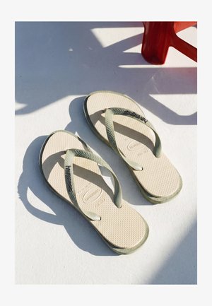 Paire de tongs beige avec des brides texturées et des semelles posées sur une surface blanche sous le soleil, ombre partielle d'une chaise rouge à proximité.
