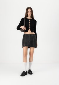 Haut noir côtelé à manches longues avec des détails de boutons, associé à une mini-jupe en cuir noir ajustée. Le modèle porte des chaussures noires et des chaussettes blanches.